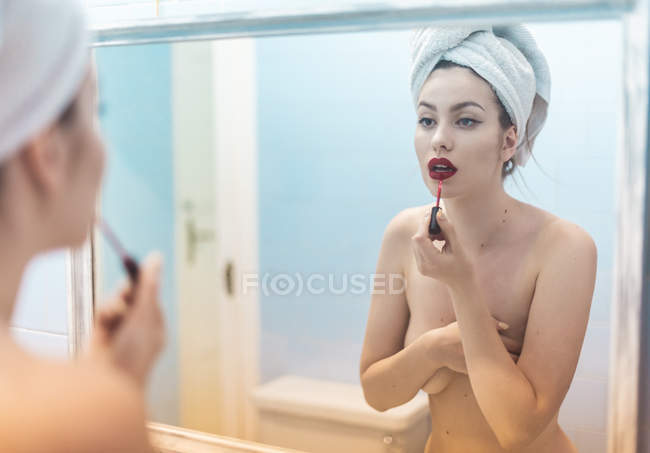 Young topless woman with makeup and towel on head standing in front of mirror in bathroom — Stock Photo