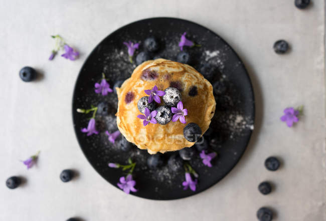 Stack of appetizing tasty crumpets with blueberries and purple flowers on black plate — Stock Photo