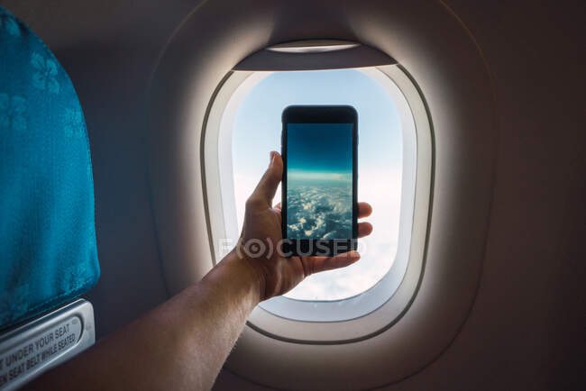 Crop hand holding smartphone and taking picture of infinite white clouds behind window of plane — Stock Photo