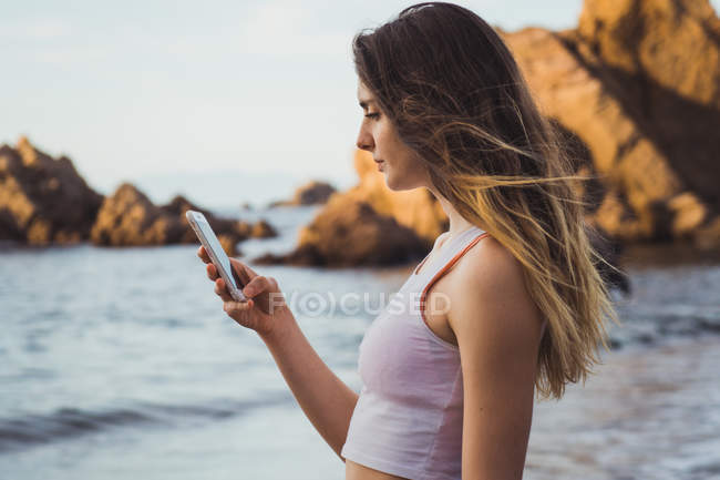 Frau in Sportkleidung steht am Meer und surft im Smartphone — Stockfoto
