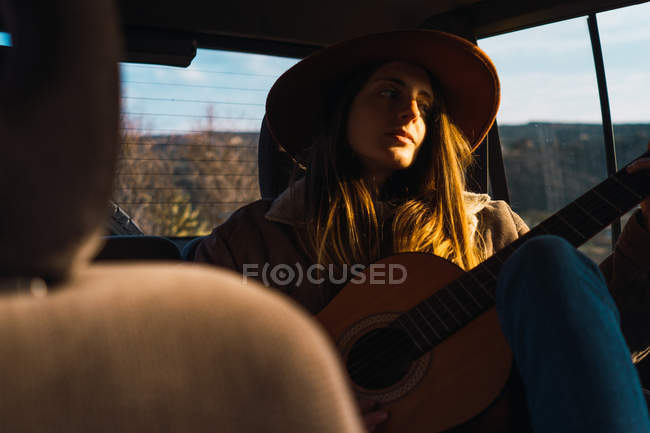 Mujer pensativa en sombrero con la guitarra sentado en el coche en la naturaleza - foto de stock