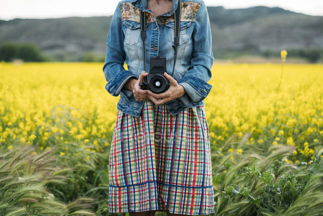 Mulher em vestido colorido e casaco de ganga segurando dispositivo de foto na natureza — Fotografia de Stock