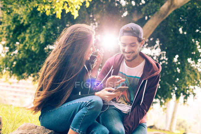 Rire jeune couple assis avec smartphone dans le parc — Photo de stock