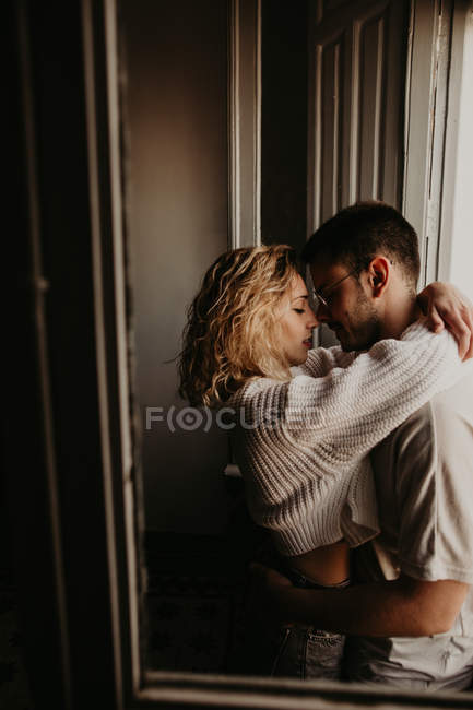 Casal afetuoso abraçando na janela em casa — Fotografia de Stock