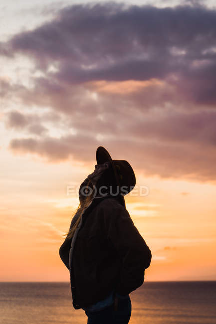 Thoughtful woman in hat standing at sea at sunset — Stock Photo