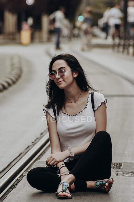 Young hipster girl in sandals and sunglasses sitting with backpack on paved tramway road in city — Stock Photo