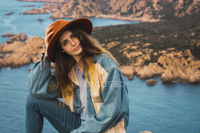 Portrait of young woman in hat sitting on rock at ocean — Stock Photo