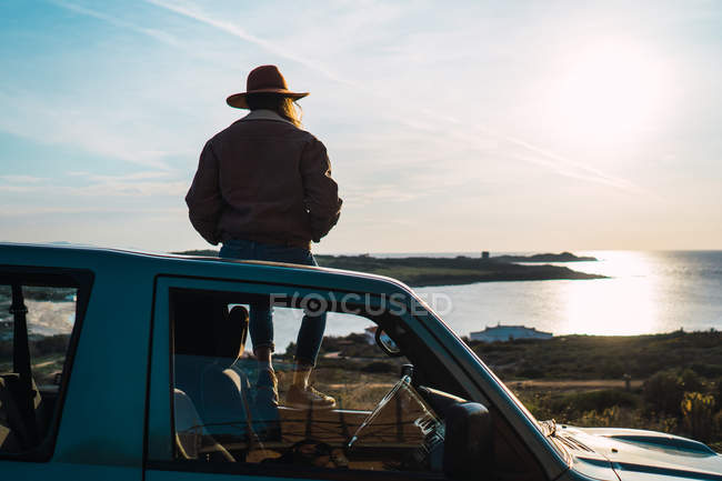 Вид сзади Женщина, сидящая на крыше автомобиля на берегу моря — стоковое фото