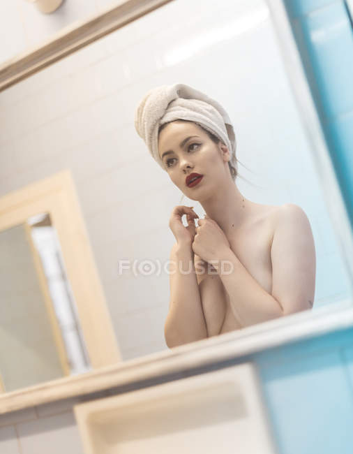 Jeune femme topless avec maquillage et serviette sur la tête debout devant le miroir dans la salle de bain — Photo de stock