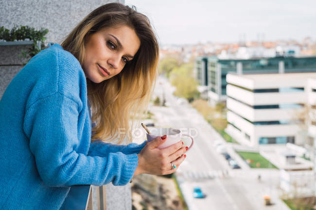Молодая женщина в синем свитере держит чашку кофе и смотрит в камеру, стоя на балконе — стоковое фото