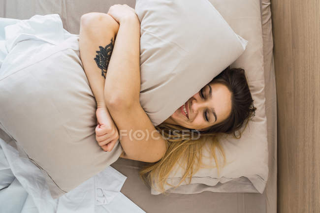 Очаровательная молодая женщина с татуировкой, счастливо лежащая на кровати и обнимающая подушку — стоковое фото
