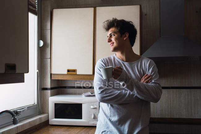 Веселий молодий чоловік стоїть з чашкою кави і дивиться на вікно на кухні — стокове фото