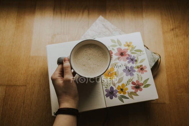 Crop hand holding cup of warm drink on book — Stock Photo