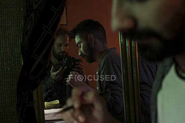 Reflection of photographer taking shots of adult man sitting and drawing indoors. — Stock Photo
