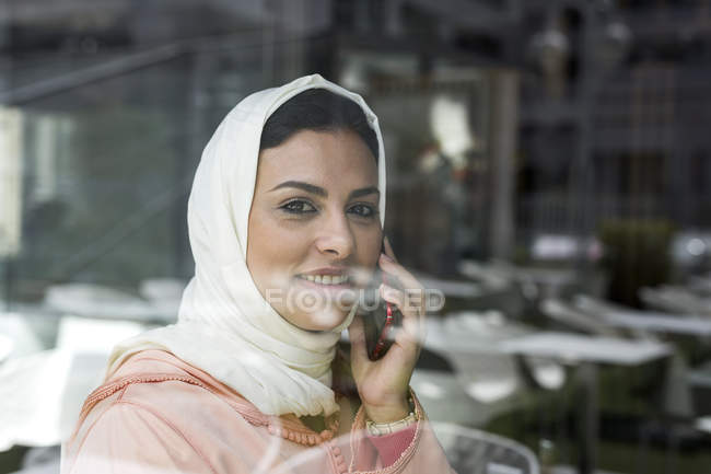 Moroccan woman with hijab and traditional Arabic dress talking on phone behind window pane — Stock Photo