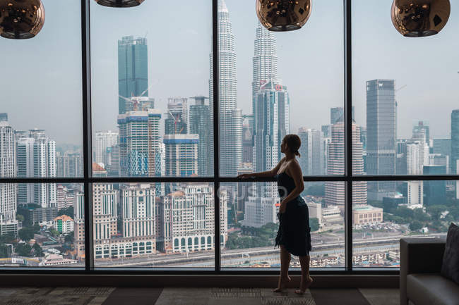 Woman standing at window in apartment and looking at skyscrapers — Stock Photo