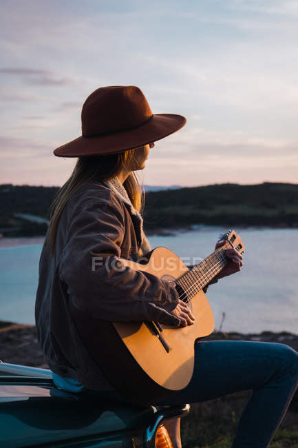 Femme assise sur le toit de la voiture et jouant de la guitare sur la côte au coucher du soleil — Photo de stock