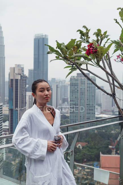 Woman in bathrobe standing at pool next to blooming tree with modern skyscrapers on background — Stock Photo