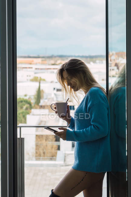 Sensual young woman in sweater holding cup of coffee and surfing smartphone in doorway of balcony — Stock Photo