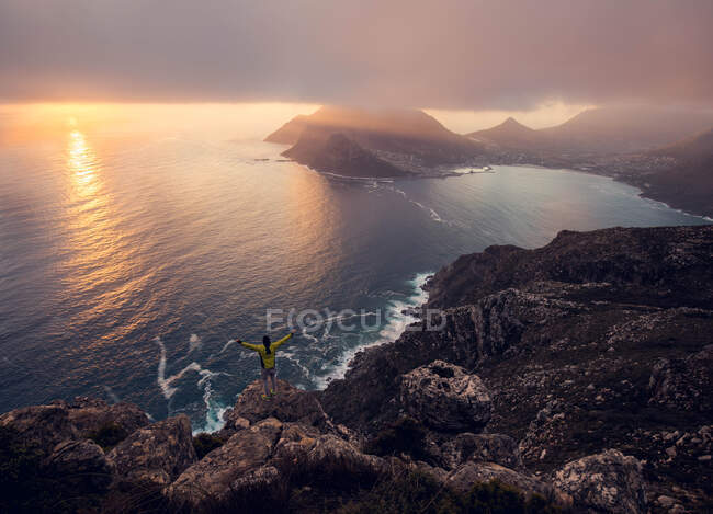 Back view of tourist standing with hands apart on rock at the ocean in sunset lights. — Foto stock