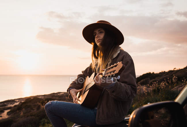 Женщина, сидящая на машине и играющая на гитаре на берегу на закате — стоковое фото