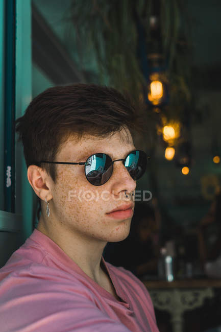 Elegante giovane uomo con piercing e orecchini che indossano occhiali da sole alla moda seduto nel bar — Foto stock