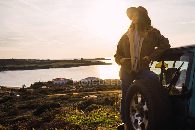Thoughtful woman leaning on car on coast at sunset — Stock Photo