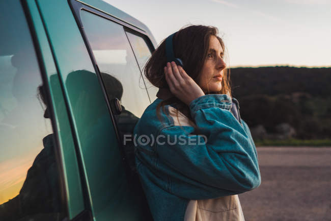 Стомлена жінка в навушниках спирається на машину — стокове фото