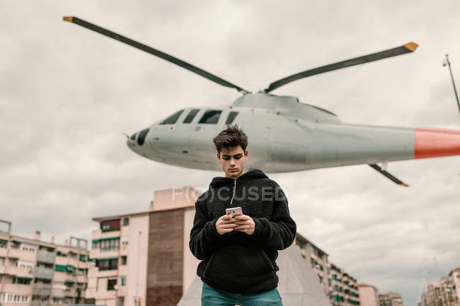 Красивий молодий чоловік стоїть біля пам'ятника вертольоту в місті і використовує смартфон — стокове фото