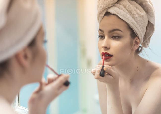 Молодая женщина топлесс с макияжем и полотенцем на голове нанося помаду перед зеркалом в ванной комнате — стоковое фото