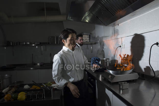 Cozinhar fazendo um flambe na cozinha do restaurante com colega no fundo — Fotografia de Stock
