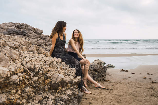 Mujer y adolescente chica sentada en la roca en la playa y hablando - foto de stock