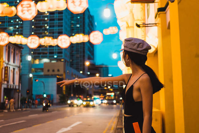 Stylish young Asian woman standing on street and catching cab in evening — Stock Photo