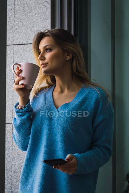 Pensive blonde woman holding smartphone and coffee cup at window — Stock Photo