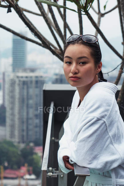 Женщина в халате стоит на балконе с видом на город и смотрит в камеру — стоковое фото