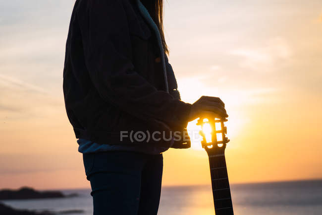 Woman in jacket standing with guitar at seaside at sunset — Stock Photo