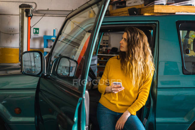 Woman using smartphone while leaning on car in garage — Stock Photo