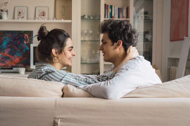 Happy young man and woman looking at each other and sitting on couch at home — Stock Photo