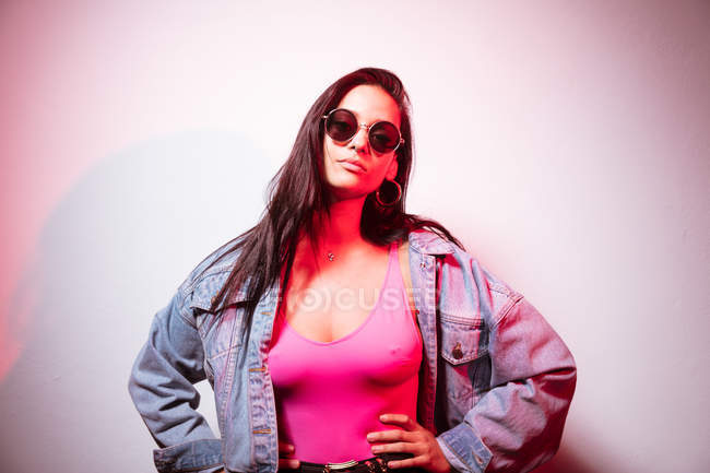Pretty young woman in sunglasses standing at white wall with hands on heap — Stock Photo
