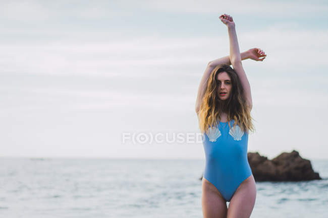 Portrait of young woman in swimsuit standing on beach with raised arms — Stock Photo