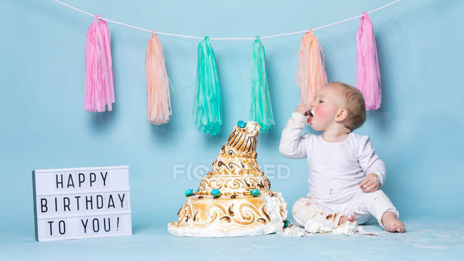 Niedlicher kleiner Junge sitzt auf Geburtstagstorte und isst auf blauem Hintergrund — Stockfoto
