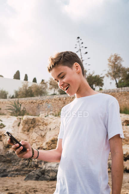 Menino com o smartphone sorrindo enquanto está de pé na costa rochosa — Fotografia de Stock