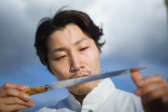 Chef japonés comprobando cuchillo delante del cielo azul - foto de stock
