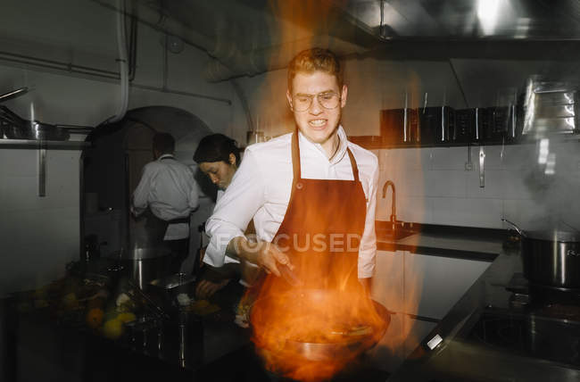 Cozinhar fazendo um flambe na cozinha do restaurante com colegas no fundo — Fotografia de Stock