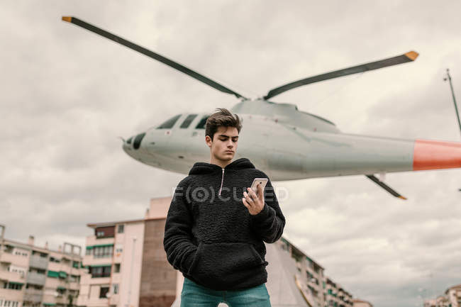 Schöner junger Mann steht am Hubschrauberdenkmal in der Stadt und benutzt Smartphone — Stockfoto