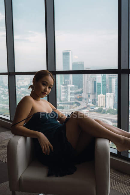 Mujer bonita en vestido negro relajante en sillón en apartamento con vista a la ciudad - foto de stock