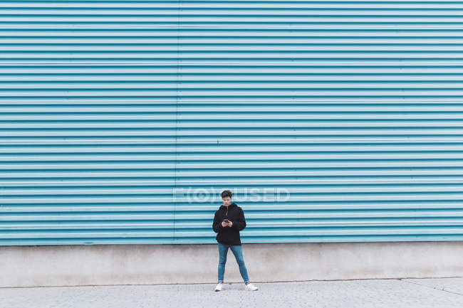 Молодой подросток, стоящий у металлической стены и использующий смартфон — стоковое фото