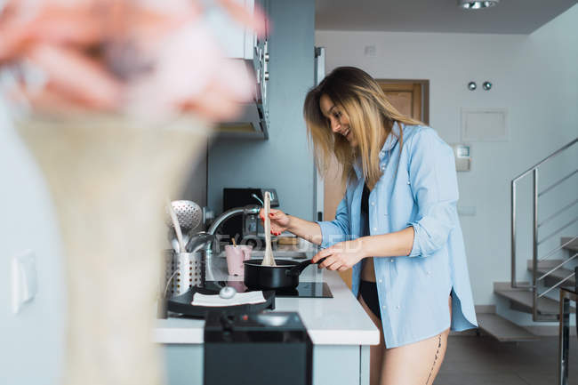 Rindo jovem mulher na camisa preparando comida na panela na cozinha — Fotografia de Stock