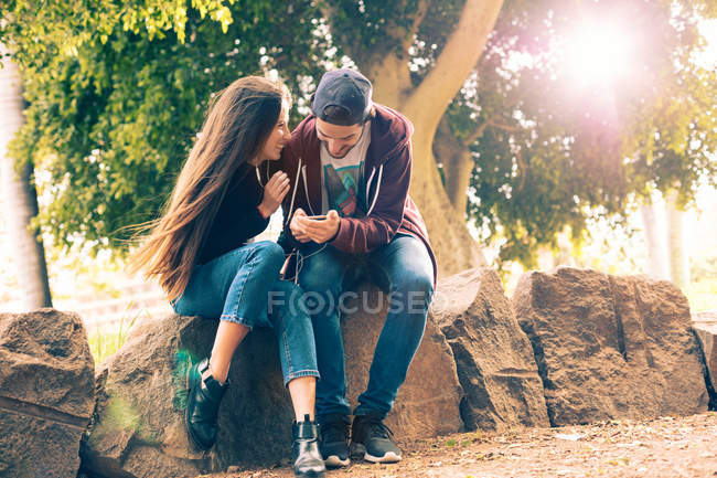 Смеющаяся молодая пара сидит на скале со смартфоном в парке — стоковое фото