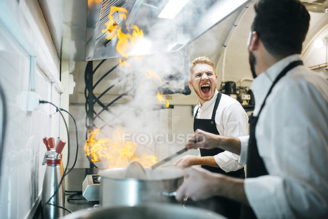 Cozinheiro animado fazendo um flambe na cozinha do restaurante com colega assistindo — Fotografia de Stock